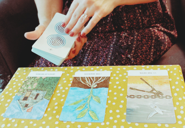 Sara is holding a tarot deck over three cards laid out in a spread on a yellow polka-dotted tablecloth.