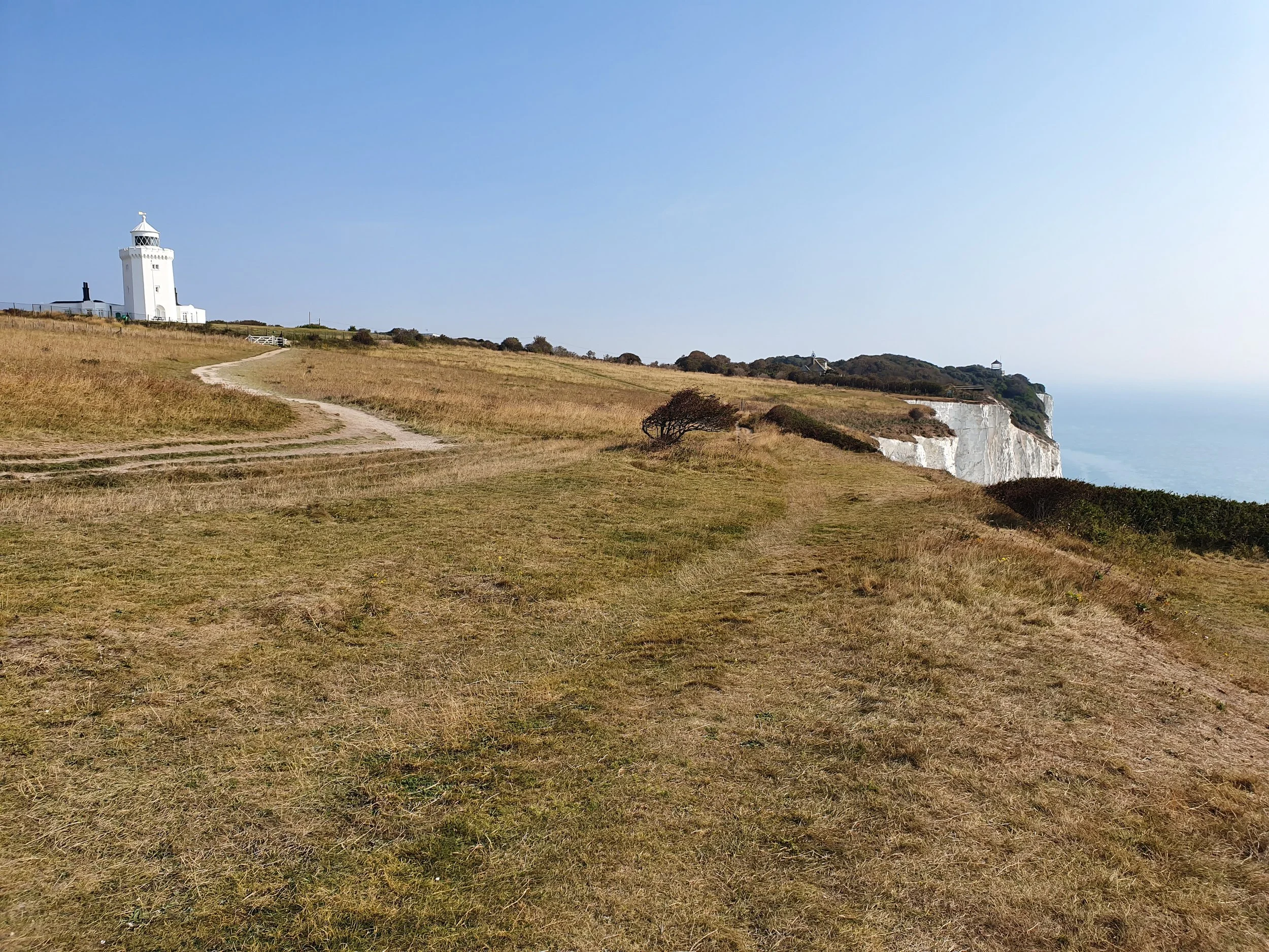 A scenic coastal landscape with a grassy hill leading to a white lighthouse on the left, and another lighthouse in the distance on a hill on the right, with cliffs and the ocean in the background under a clear blue sky.