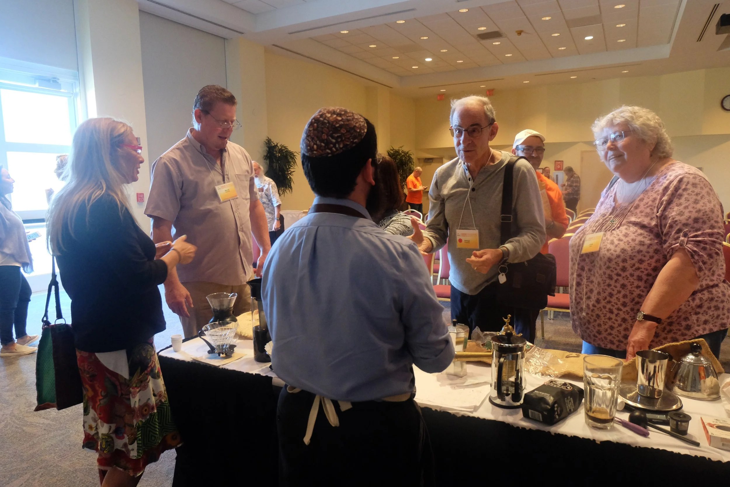 People gathering around a table at an indoor event, engaged in conversation, with various items including coffee equipment on the table.