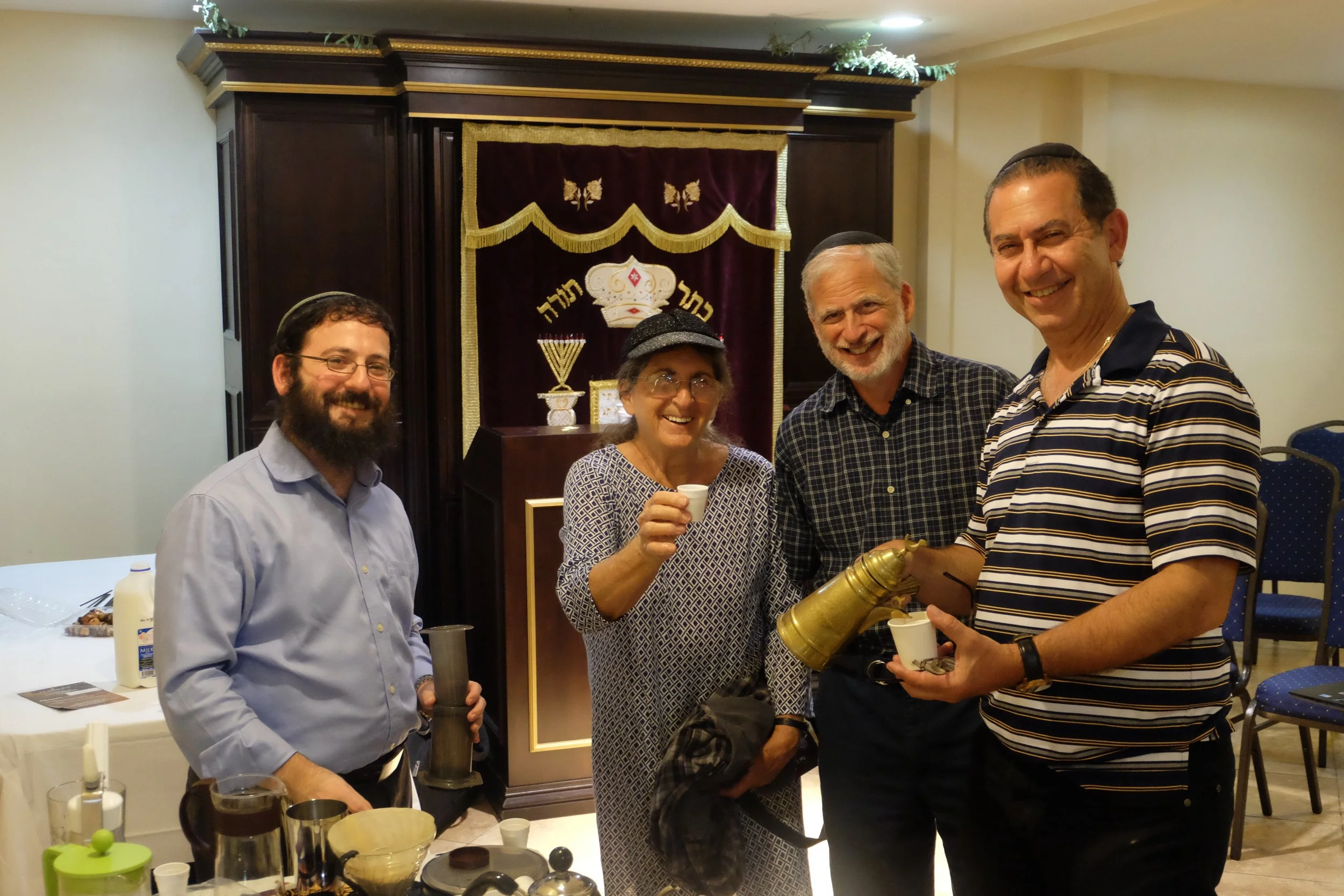 Four people smiling at a celebration or gathering, with a decorative background featuring Jewish symbols, including a crown and menorah. They are holding ceremonial items, and there are food and drinks on the table.