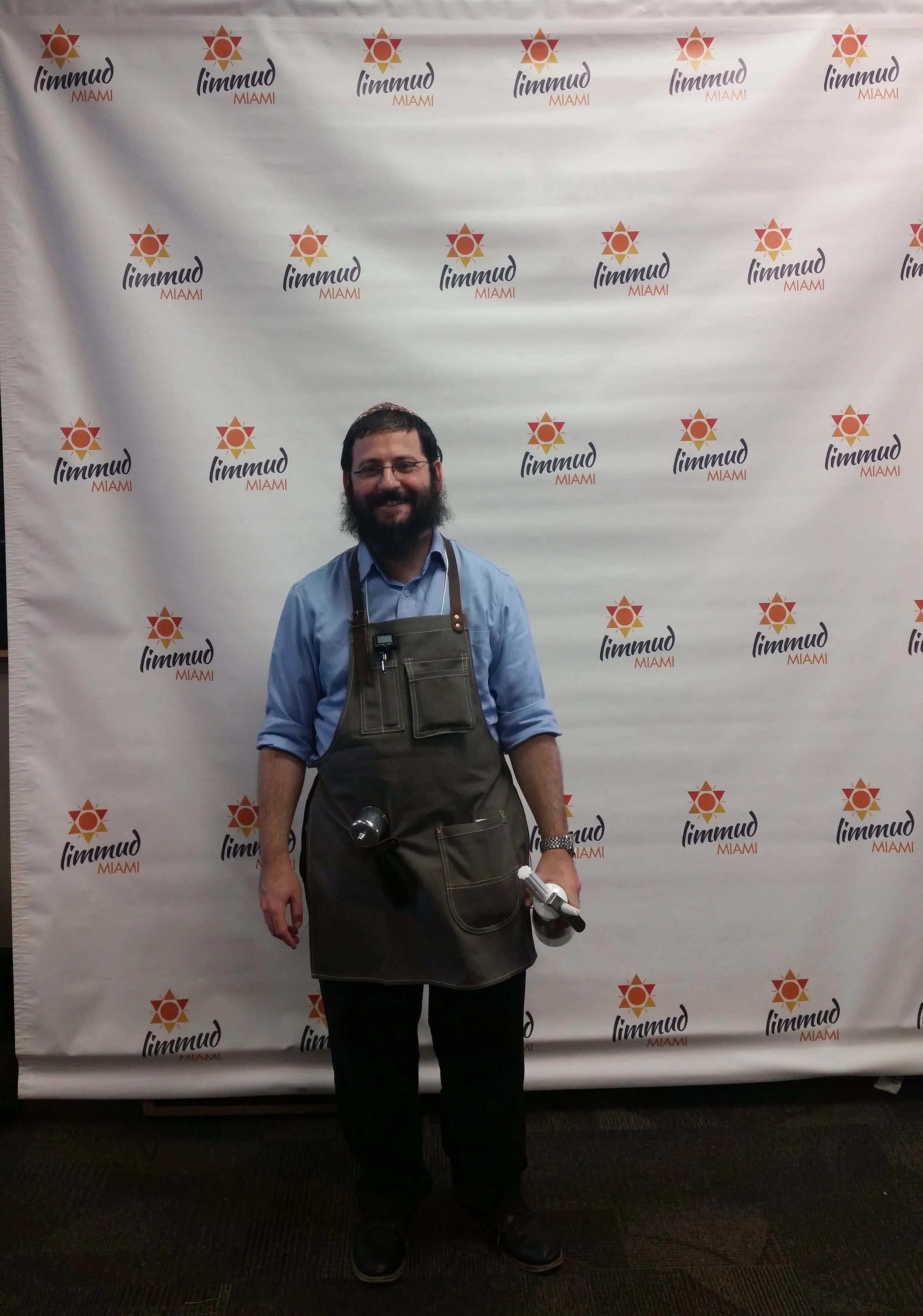 A man with glasses, a beard, wearing a blue shirt and black pants, stands in front of a backdrop with the logo and the words 'limmud MIAMI'. He is wearing an apron and holding a water bottle and a microphone in his left hand, smiling.