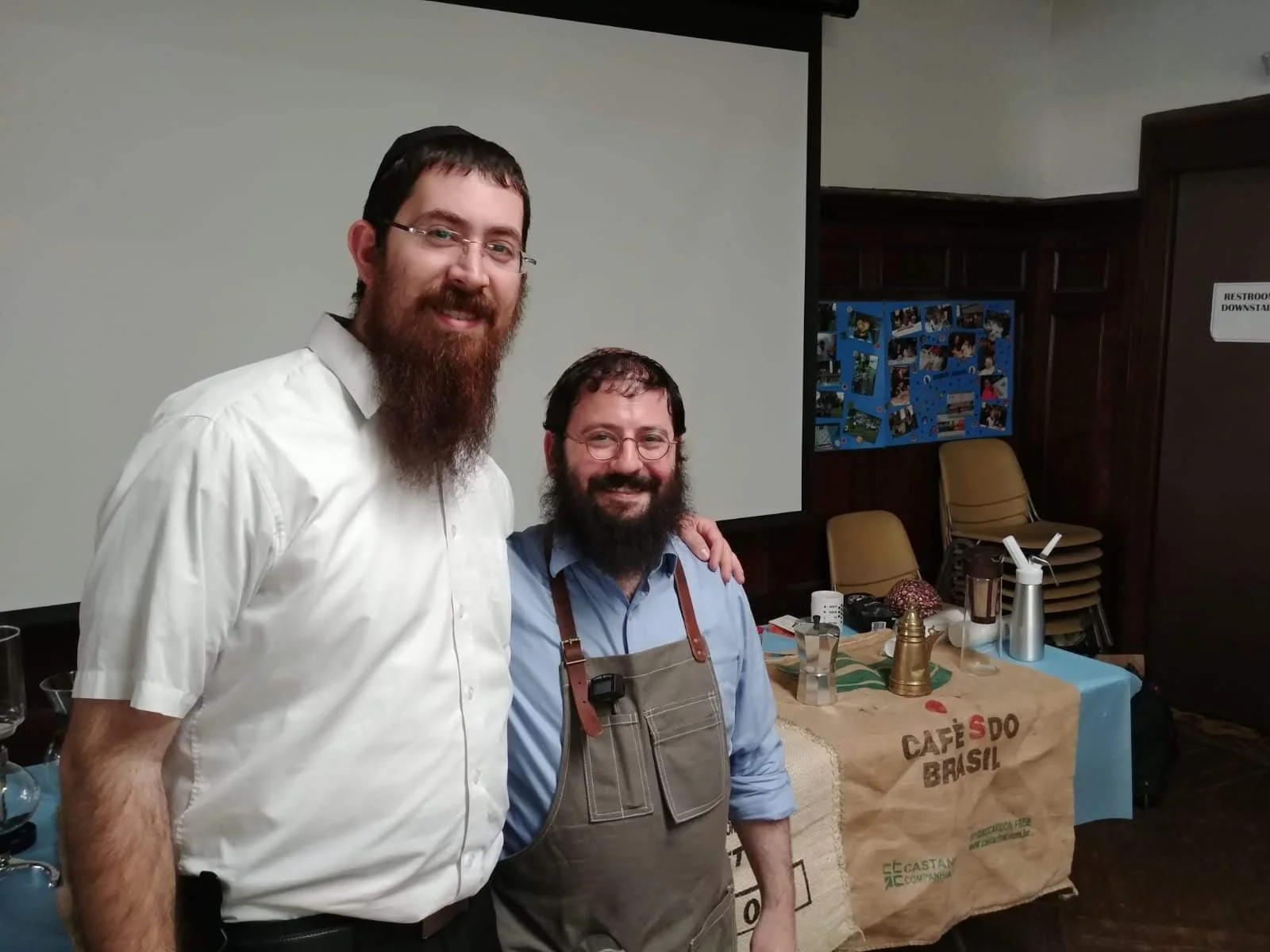 Two men with beards and glasses smiling, standing in front of a table with coffee and teapots, in a room with a large white screen and a bulletin board with photos.