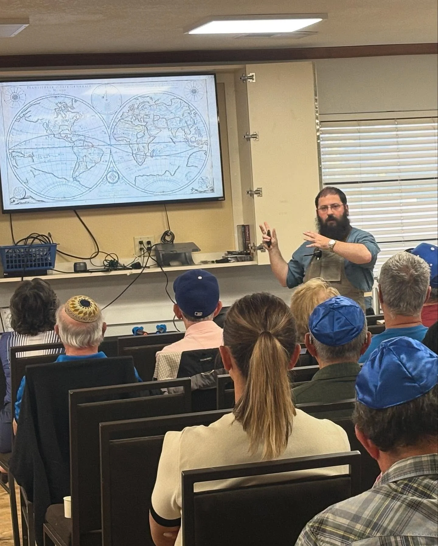 A man with a beard and glasses giving a presentation in a room filled with seated audience members. A large map of the world is projected on a screen behind him.