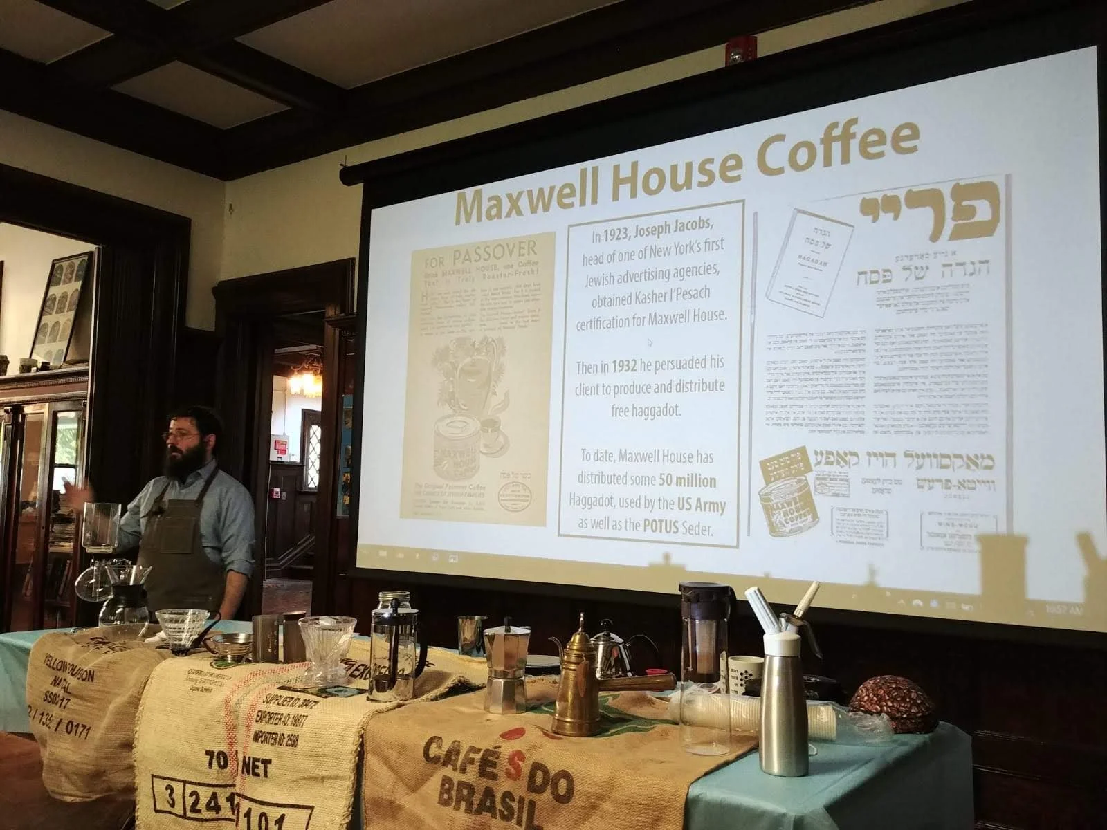 Presentation slide about Maxwell House Coffee displayed behind a man in a workshop or seminar setting, with tables covered in burlap sacks of coffee beans and coffee-making equipment in the foreground.