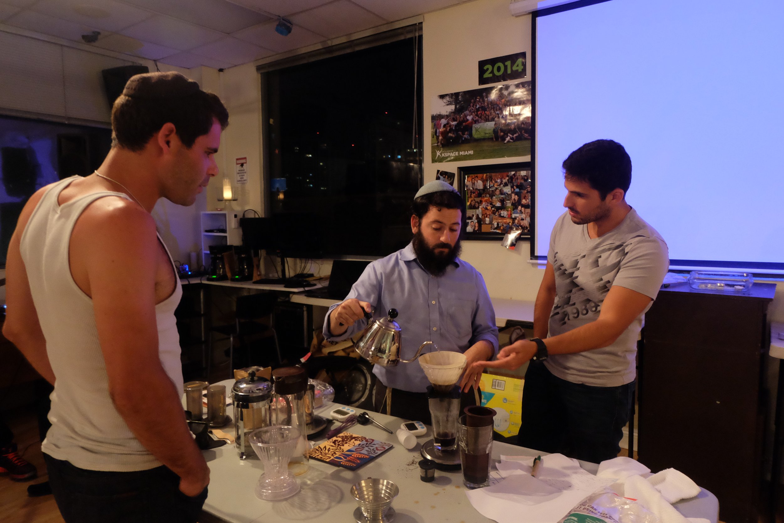 Three men are conducting a coffee brewing demonstration or workshop at a table with coffee-making equipment. One man is pouring hot water over coffee grounds in a pour-over coffee maker, while the others observe. The setting appears to be a modern indoor space with posters and photos on the wall, including a large poster labeled '2014' and a collage of photos below it.
