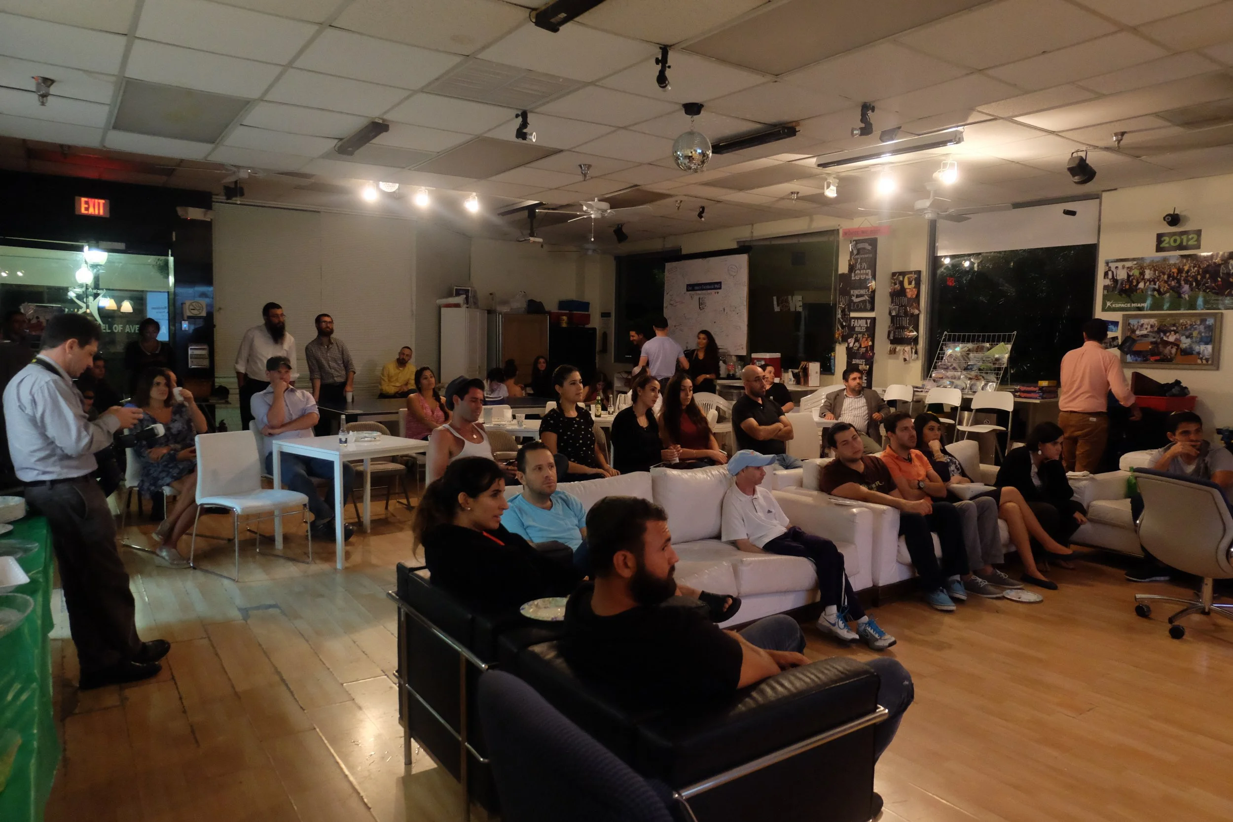People seated and standing inside a room, watching a presentation or event, with some standing near the back and a few at tables and on couches, with posters on the walls and a window showing the outdoors.