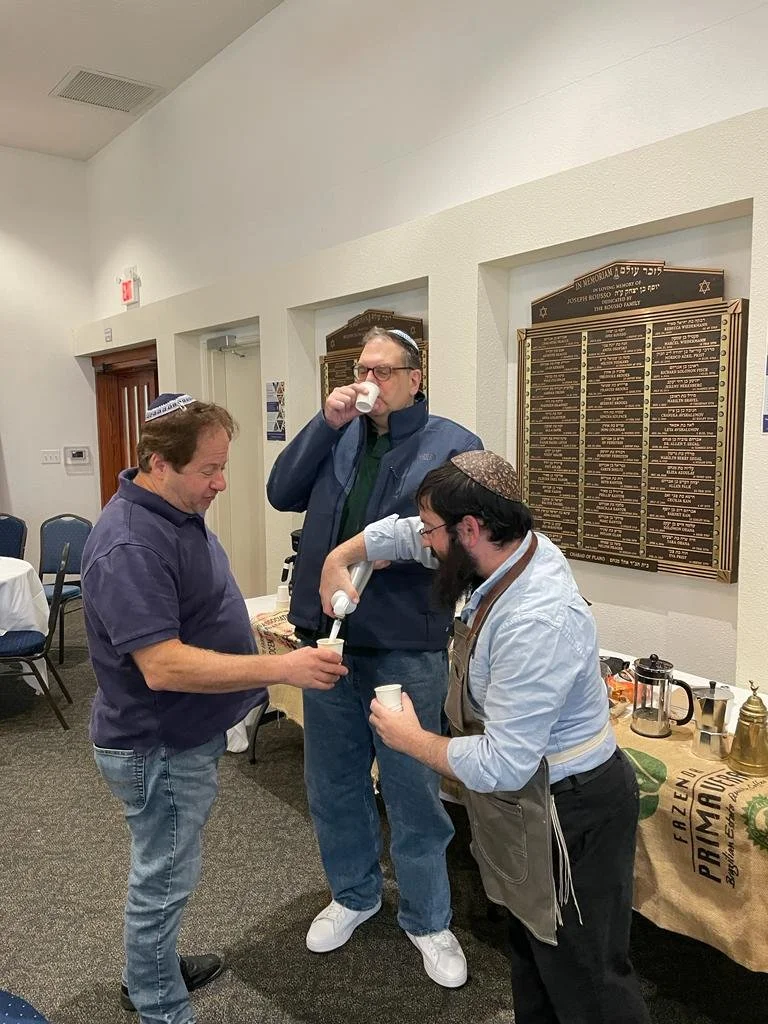Three men participating in a traditional Jewish ritual called 'Kiddush' with wine and cups in a room with a memorial plaque on the wall.