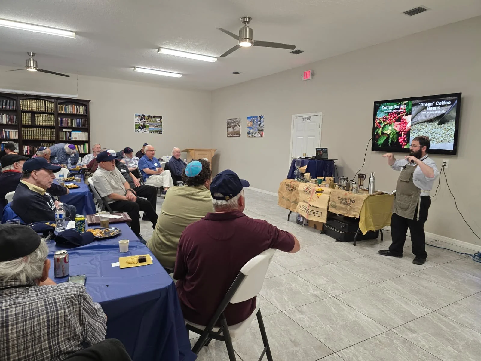 A man wearing an apron is giving a presentation about coffee to an audience sitting at tables with blue tablecloths. A large screen behind him shows images of coffee beans and berries.