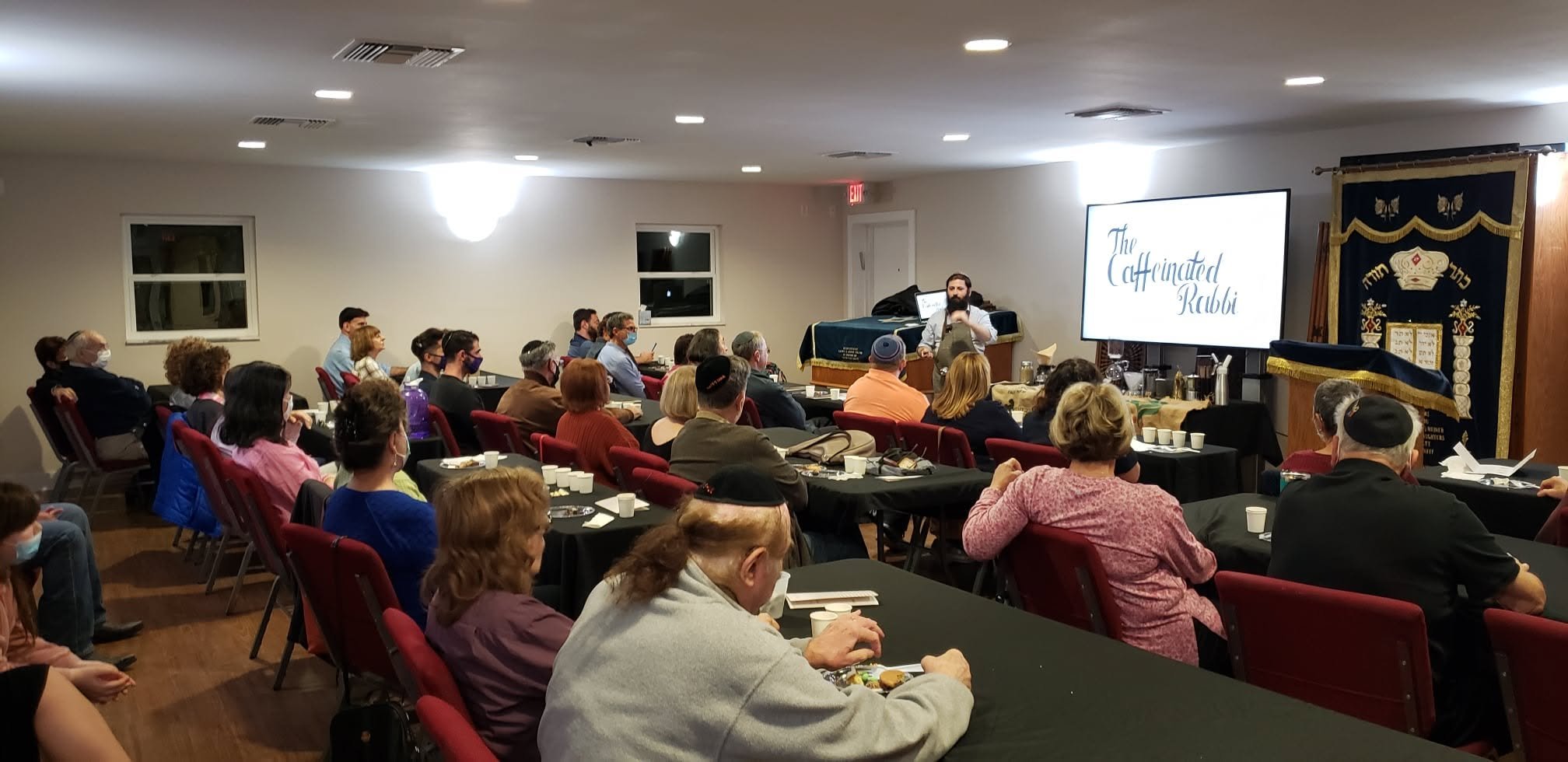 A man stands at the front of a room giving a presentation to an audience seated at tables. The screen behind him shows the words 'The Caffeinated Rubi.' The audience is diverse, with some wearing masks and religious head coverings, and some eating snacks.