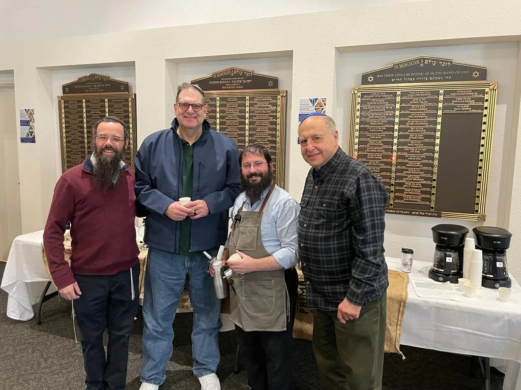Four men standing together indoors in front of memorial plaques on the wall, smiling at the camera. Two coffee makers and cups are on a table to the right.