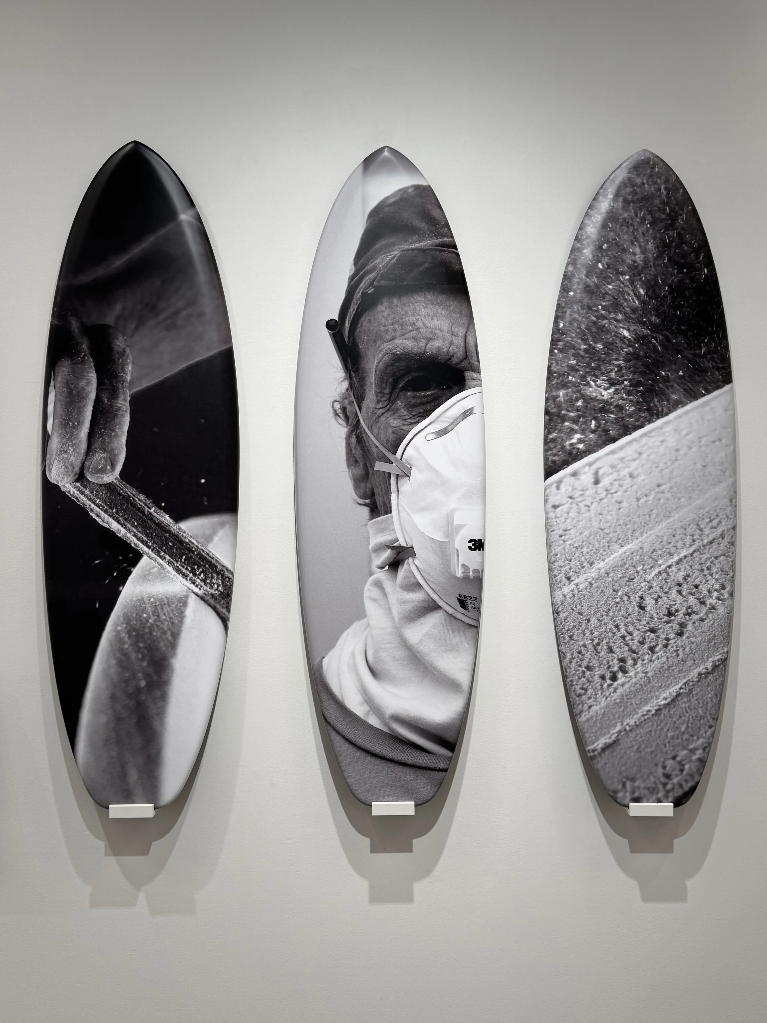 Three surfboards mounted on a white wall, featuring black-and-white photos: a close-up of a hand holding a surfboard on the left, a portrait of a woman wearing a face mask in the middle, and a sandy beach with some water on the right.