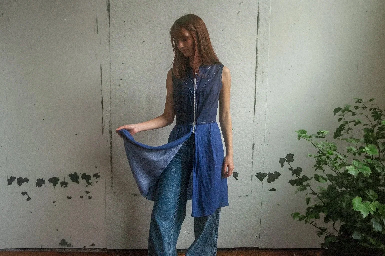 A woman with reddish-brown hair wearing a sleeveless dark blue top with a zipper, and blue jeans, standing indoors against a textured light gray wall next to a green leafy plant. She is looking down and holding the hem of her top.