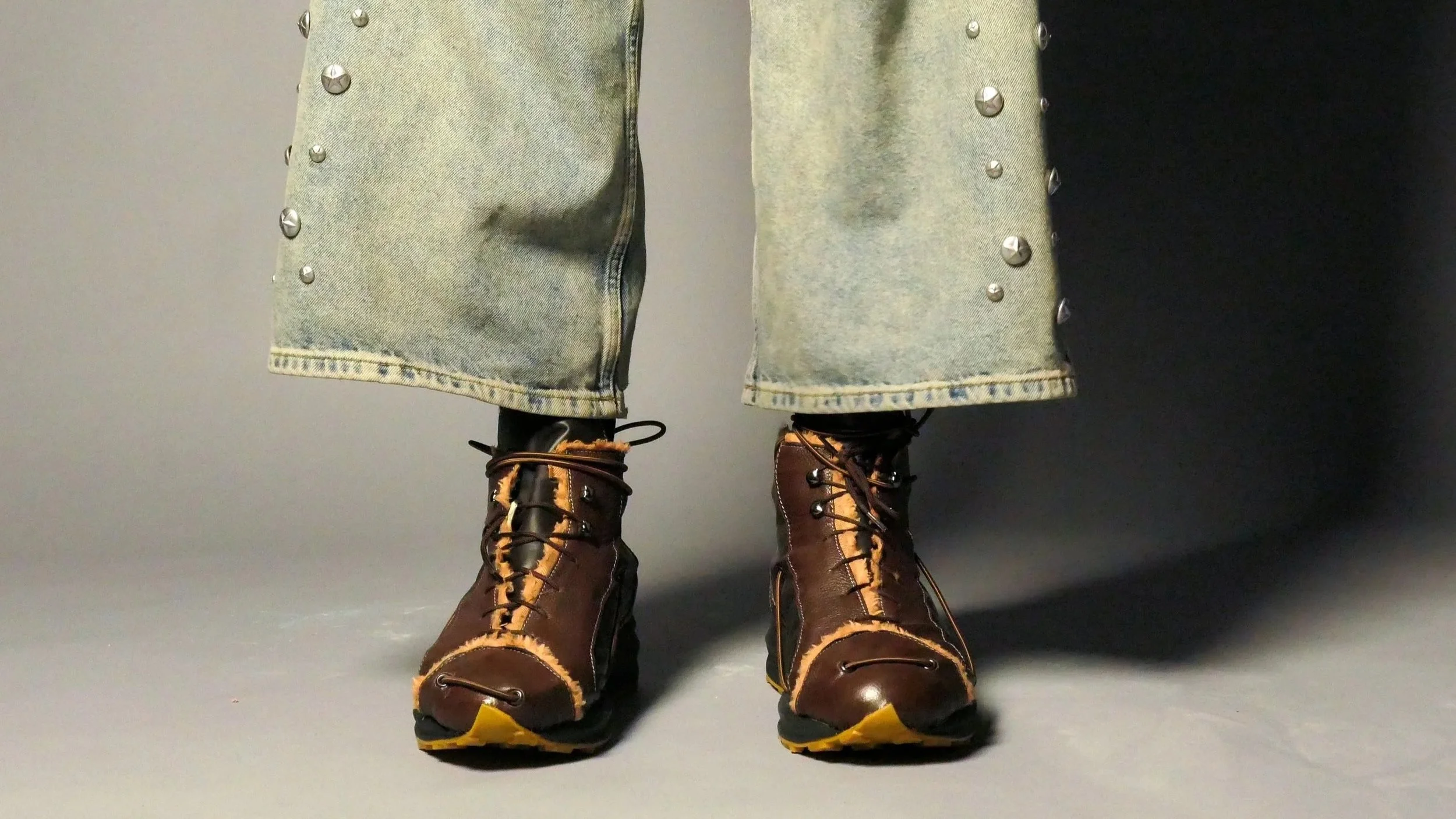 Close-up of a person's legs wearing distressed denim jeans with metal studs and brown hiking boots with yellow accents, standing on a gray surface against a gray and black background.