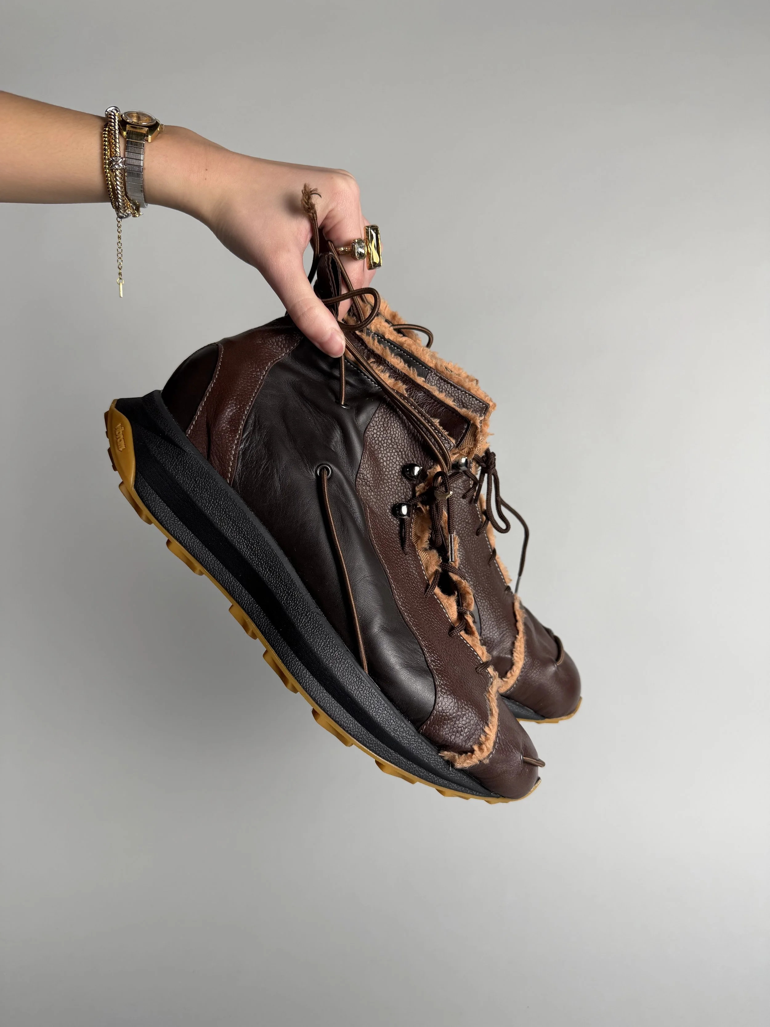 A person's hand holding a brown leather and fabric ankle boot with a rugged sole, against a plain gray background.