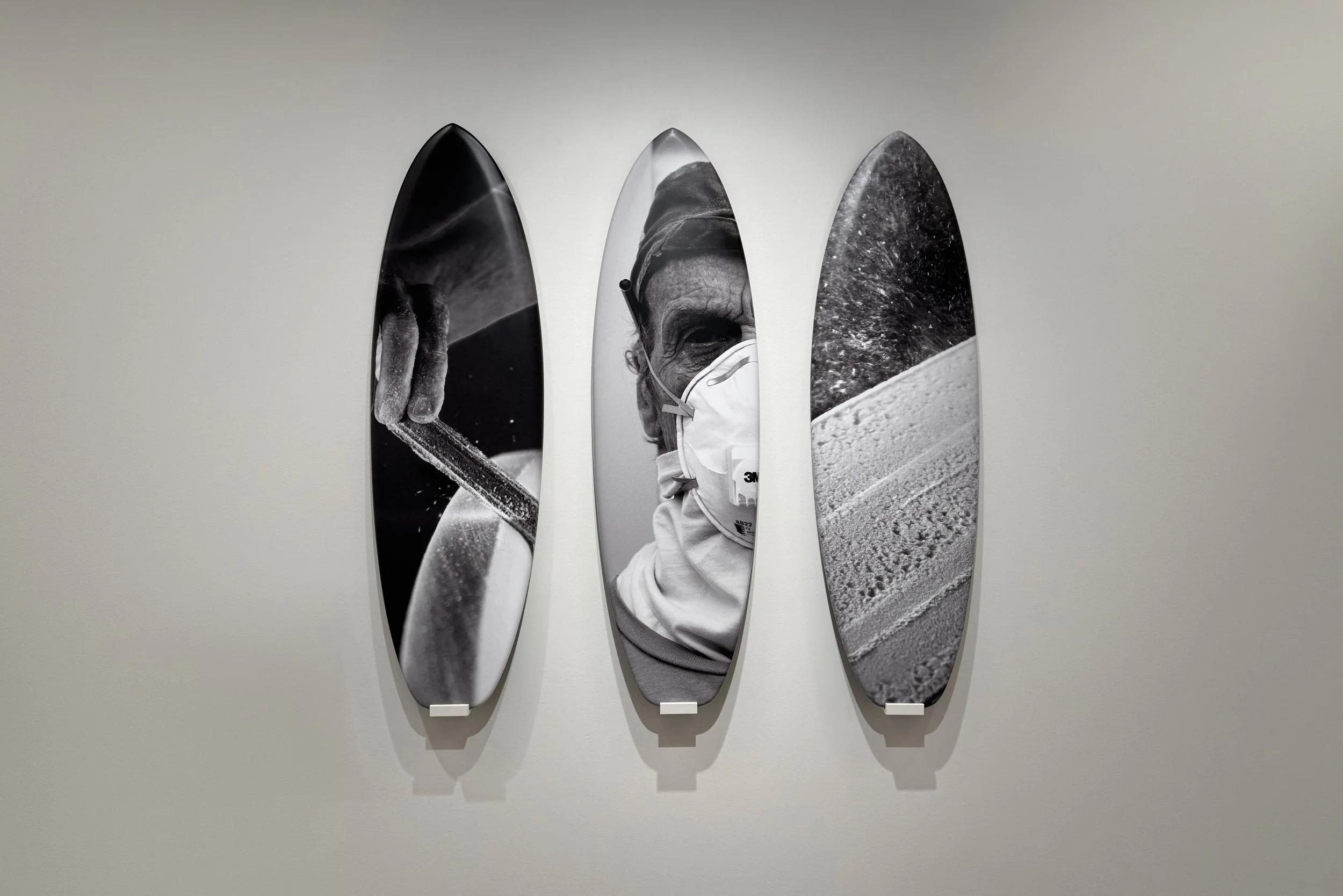 Black and white artistic wall display of vertical surfboard-shaped panels featuring close-up photographs of a scientist or medical professional wearing a face mask, holding a pipette, and working with scientific material, along with a textured surface resembling ground or sand.