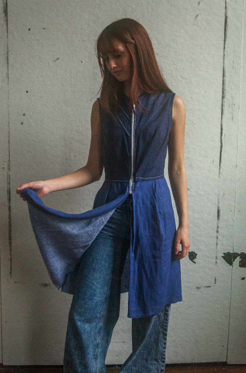 A woman with reddish-brown hair wears a sleeveless blue dress with a zipper in the front and blue jeans, standing against a plain wall.