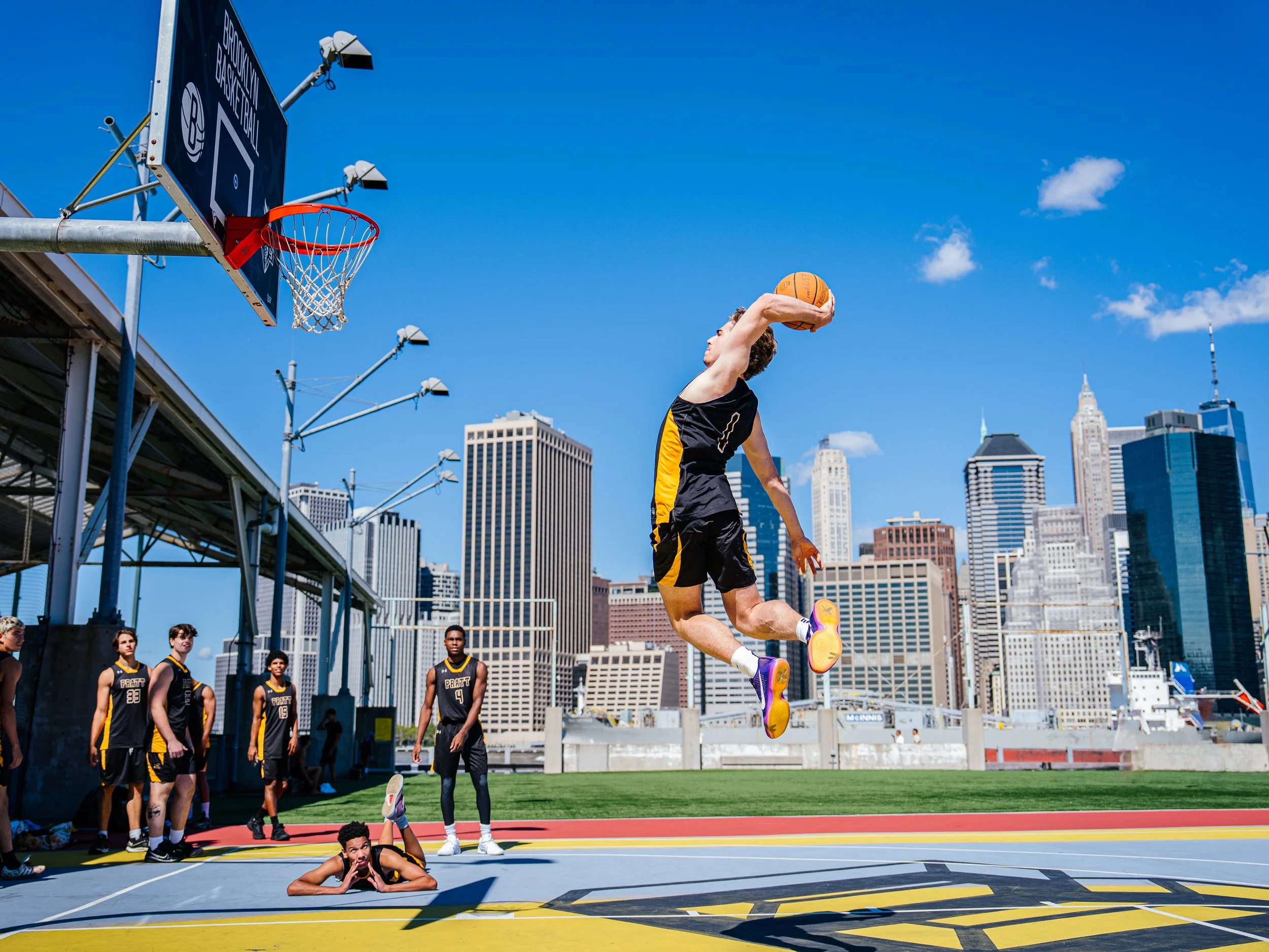 A basketball player is mid-air attempting a dunk during a game on an outdoor court in a city. Several other players are on the court, and a city skyline with tall buildings is visible in the background. The sky is clear and blue.