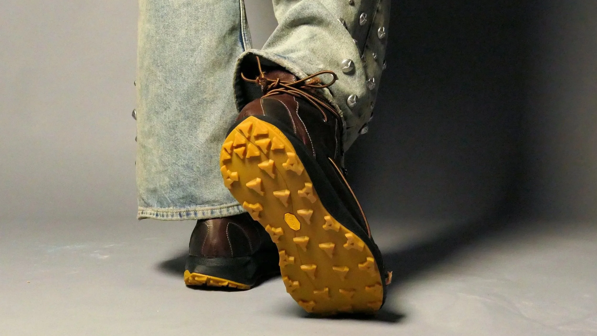 Close-up of a person's lower body showing brown hiking boots with yellow soles, black laces, and metal eyelets, khaki pants, and a light blue denim jacket with metal studs on the cuff, against a gray background.
