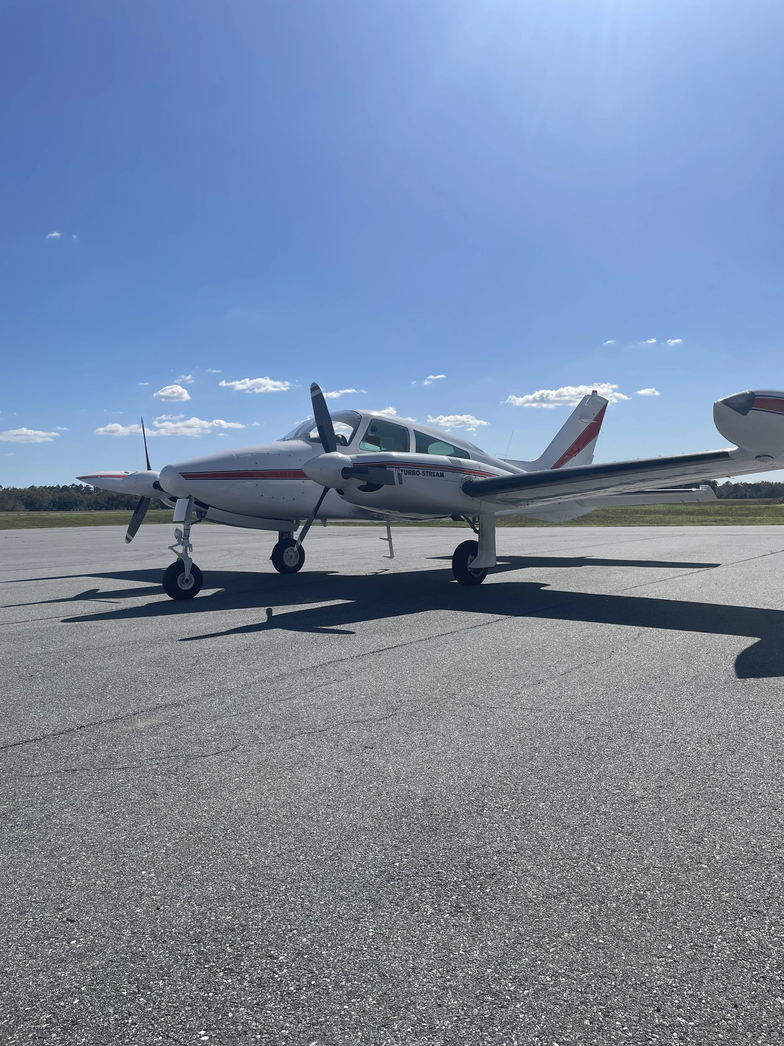 A small white airplane with a propeller, parked on a tarmac under a blue sky with some scattered clouds.