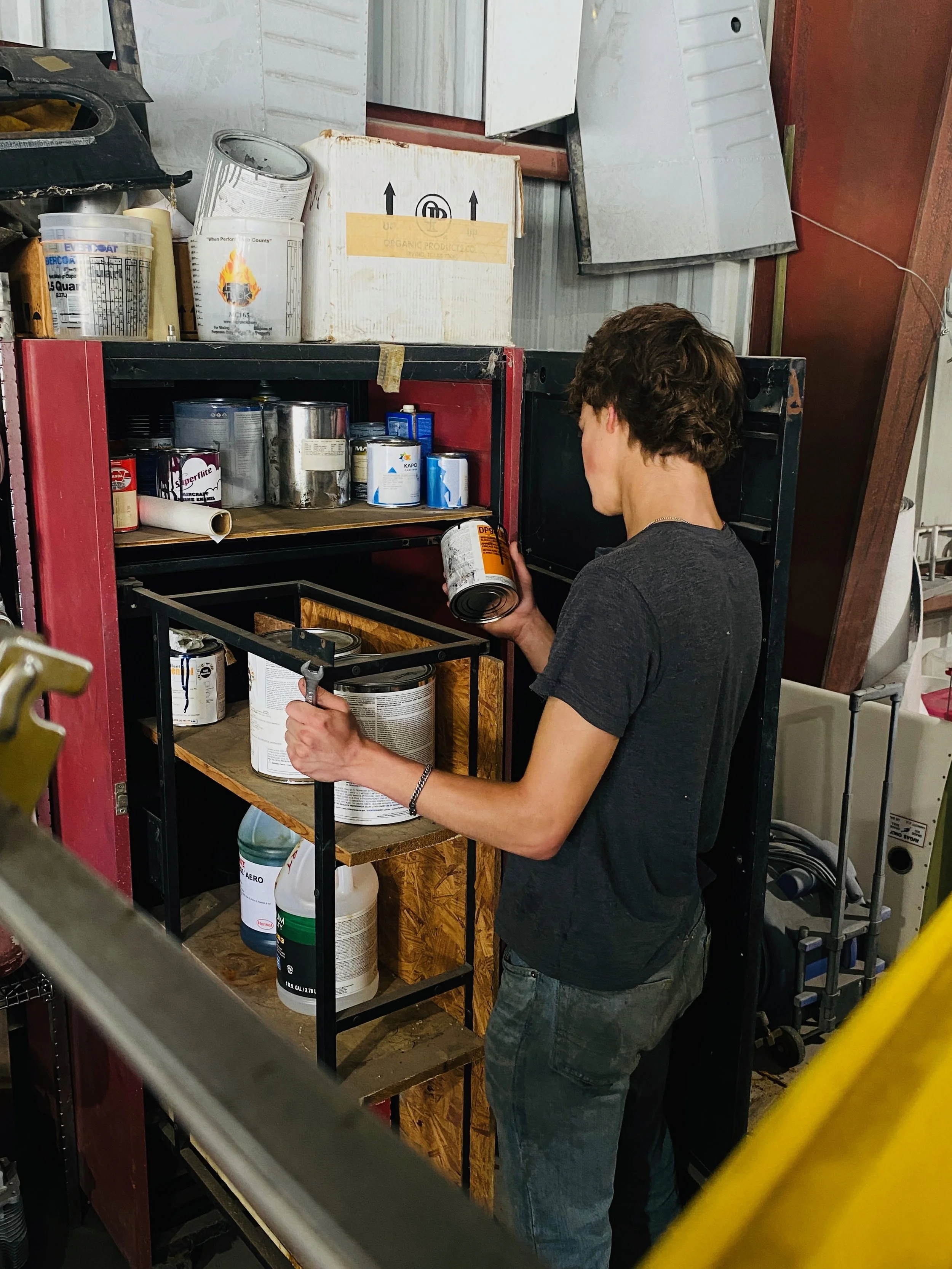 Person with short brown hair and casual clothing looking at a can of paint or other liquid in a workshop or storage room with shelves filled with various paint cans and supplies.