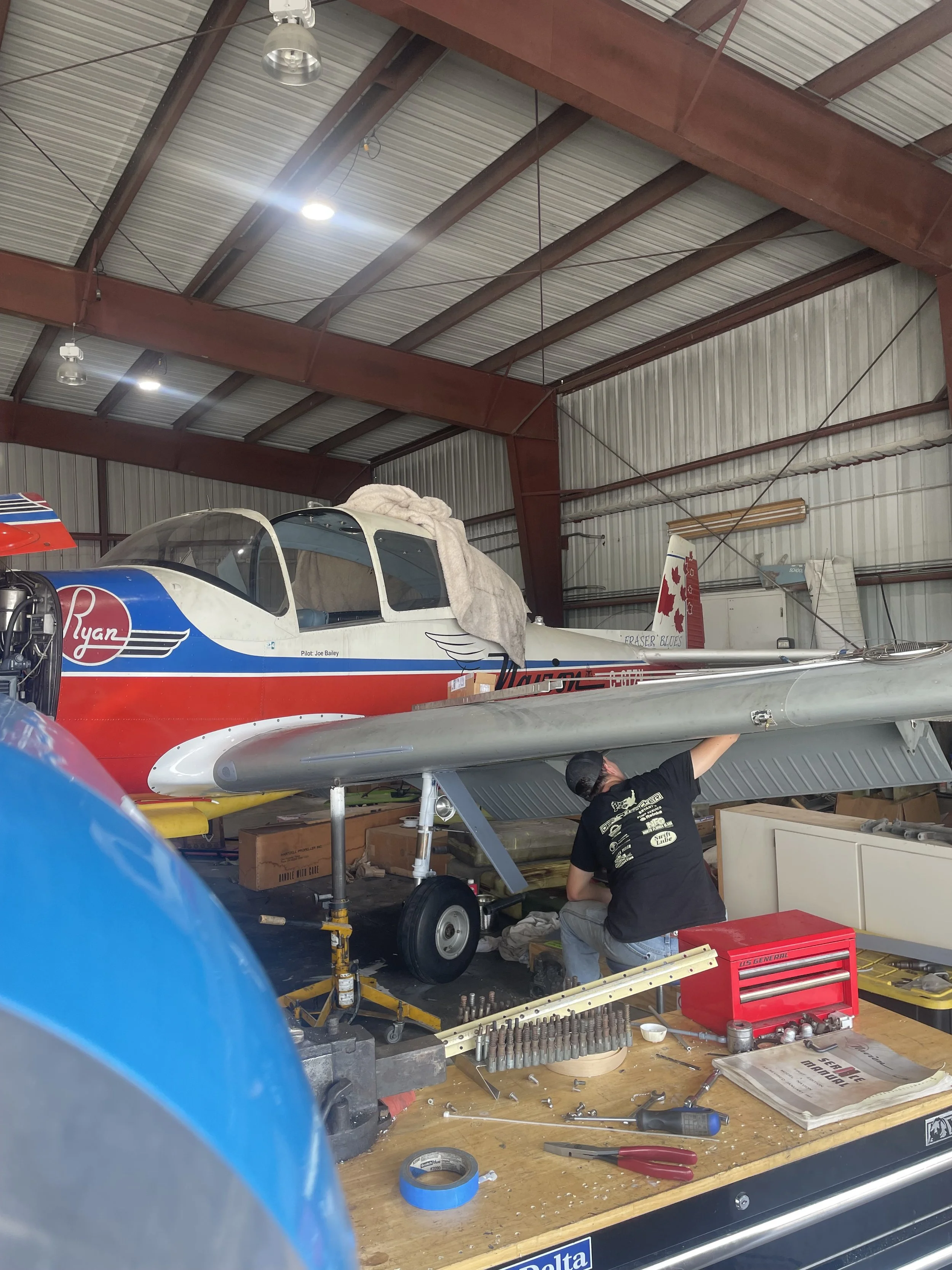 Inside an aircraft hangar, a mechanic is working under a small airplane, with tools and equipment spread out on a workbench. The hangar has a metal roof with exposed beams and lighting fixtures.