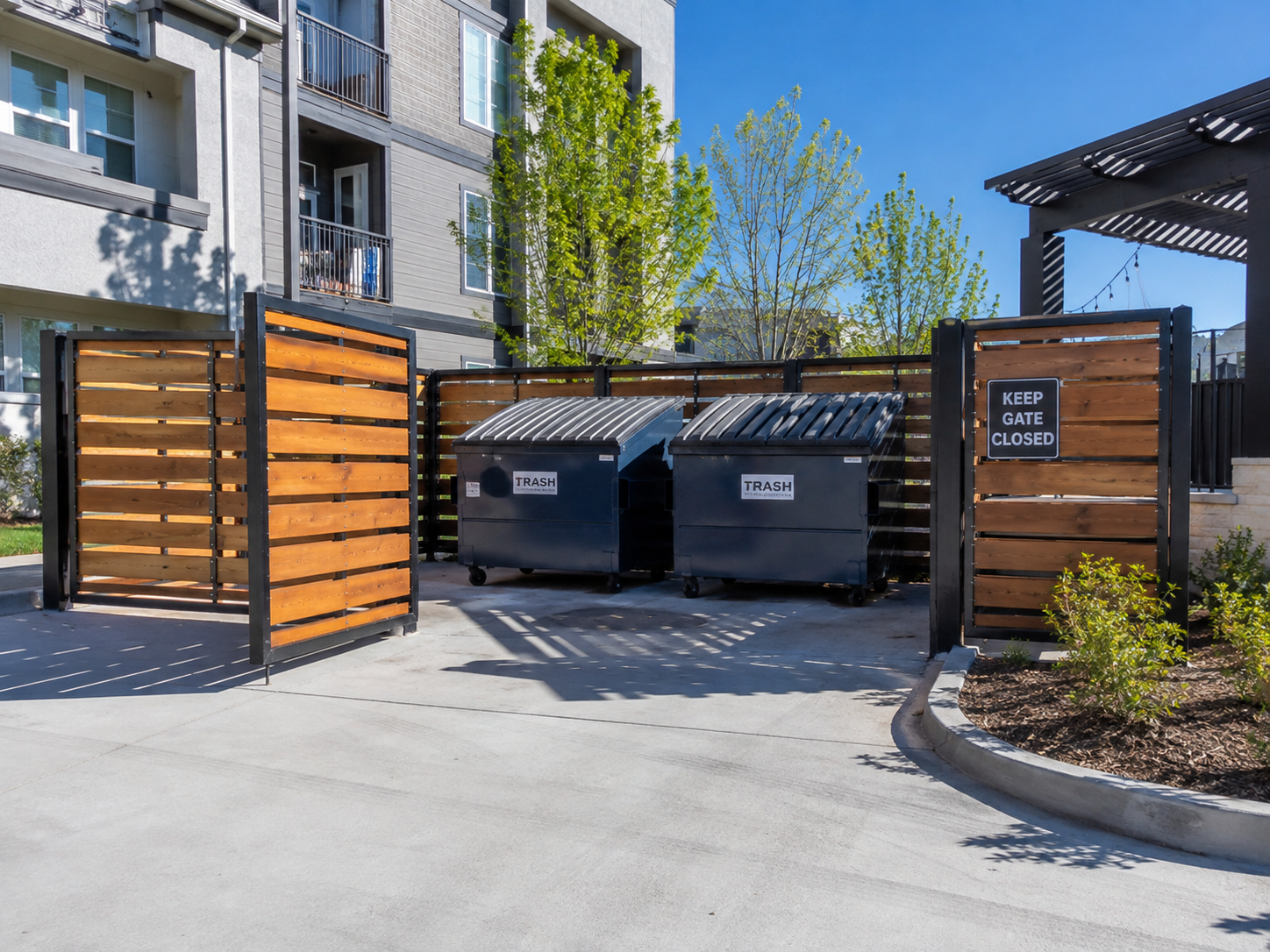 Two black trash dumpsters with labeled signs, one on each side, behind a wooden fenced gate that has very clean dumpster area. The area is part of an apartment complex with residential buildings, trees, and a clear blue sky.
