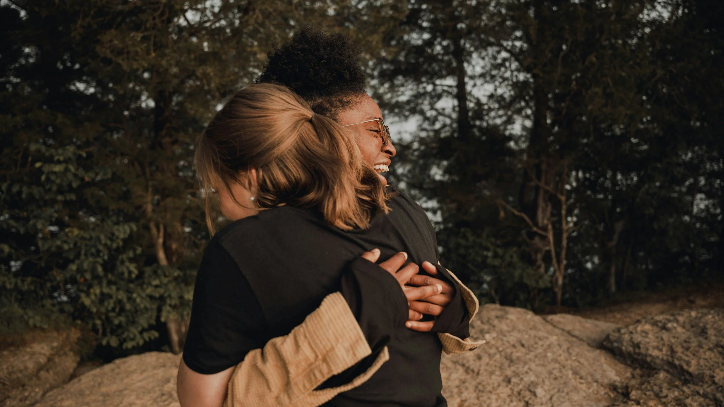 Two women hugging outdoors near trees, smiling and enjoying a moment together.