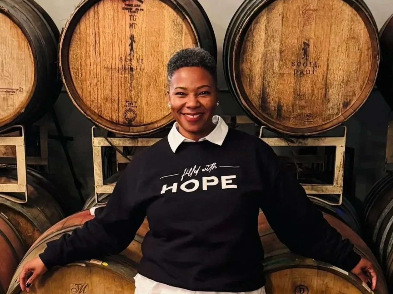 A woman smiling in a black sweatshirt with the words 'filled with HOPE' standing in front of wine barrels.