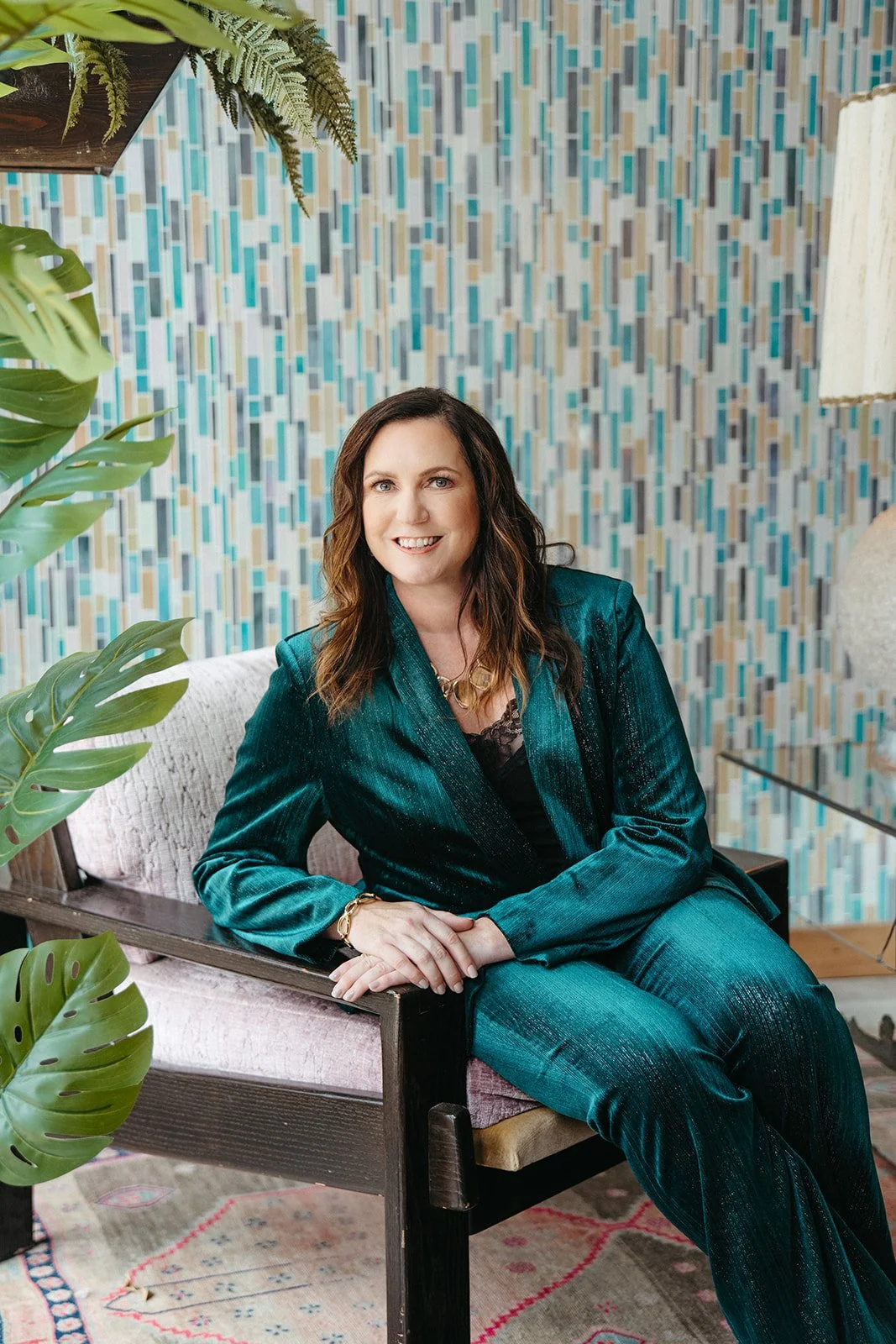 A woman with shoulder-length brown hair, wearing a teal velvet suit, sitting on a couch with a patterned blue and beige background and green plants nearby.