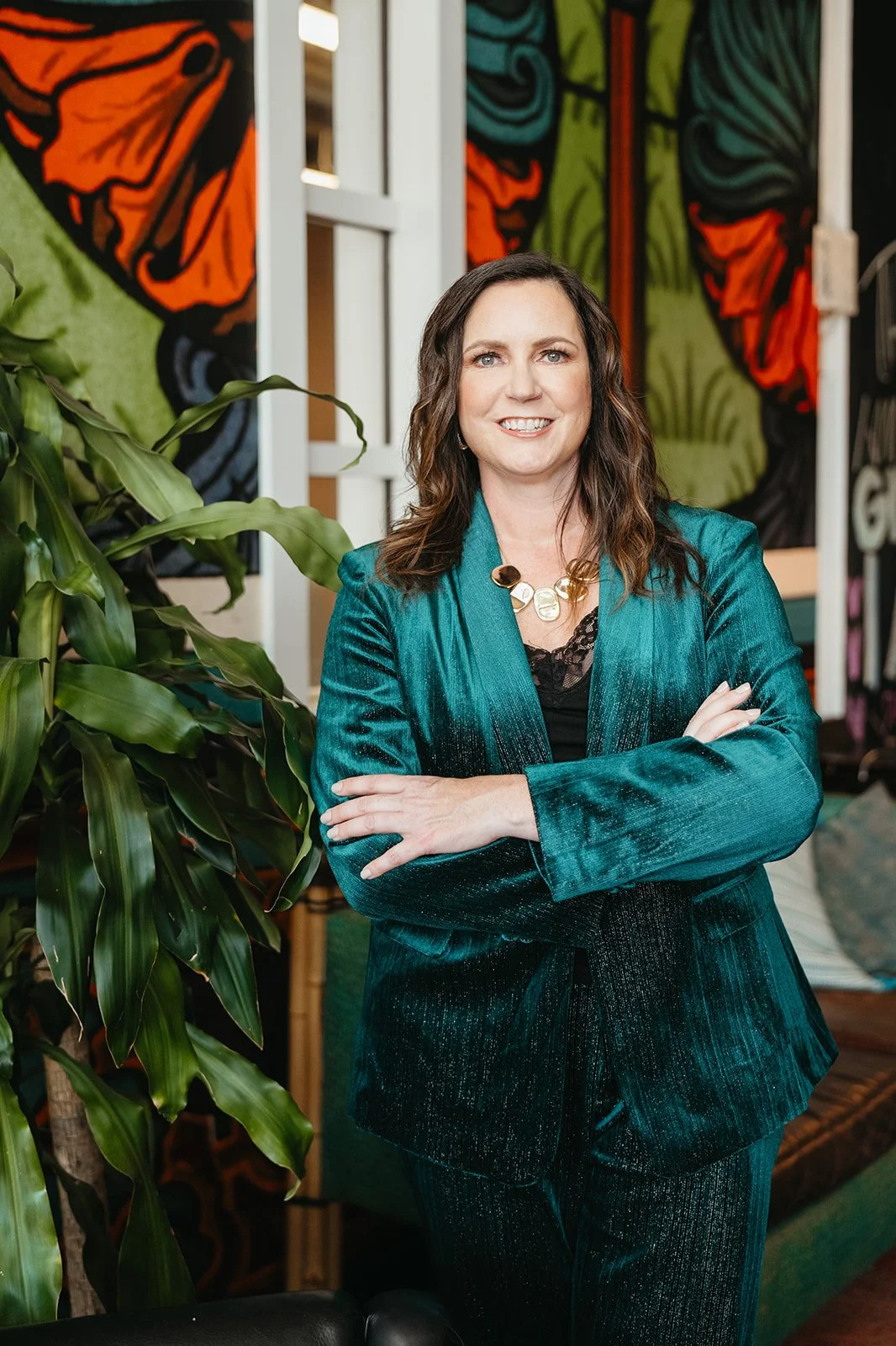 A woman with shoulder-length brown hair, wearing a teal velvet blazer and matching pants, stands with arms crossed in front of colorful abstract wall art and a large green plant.