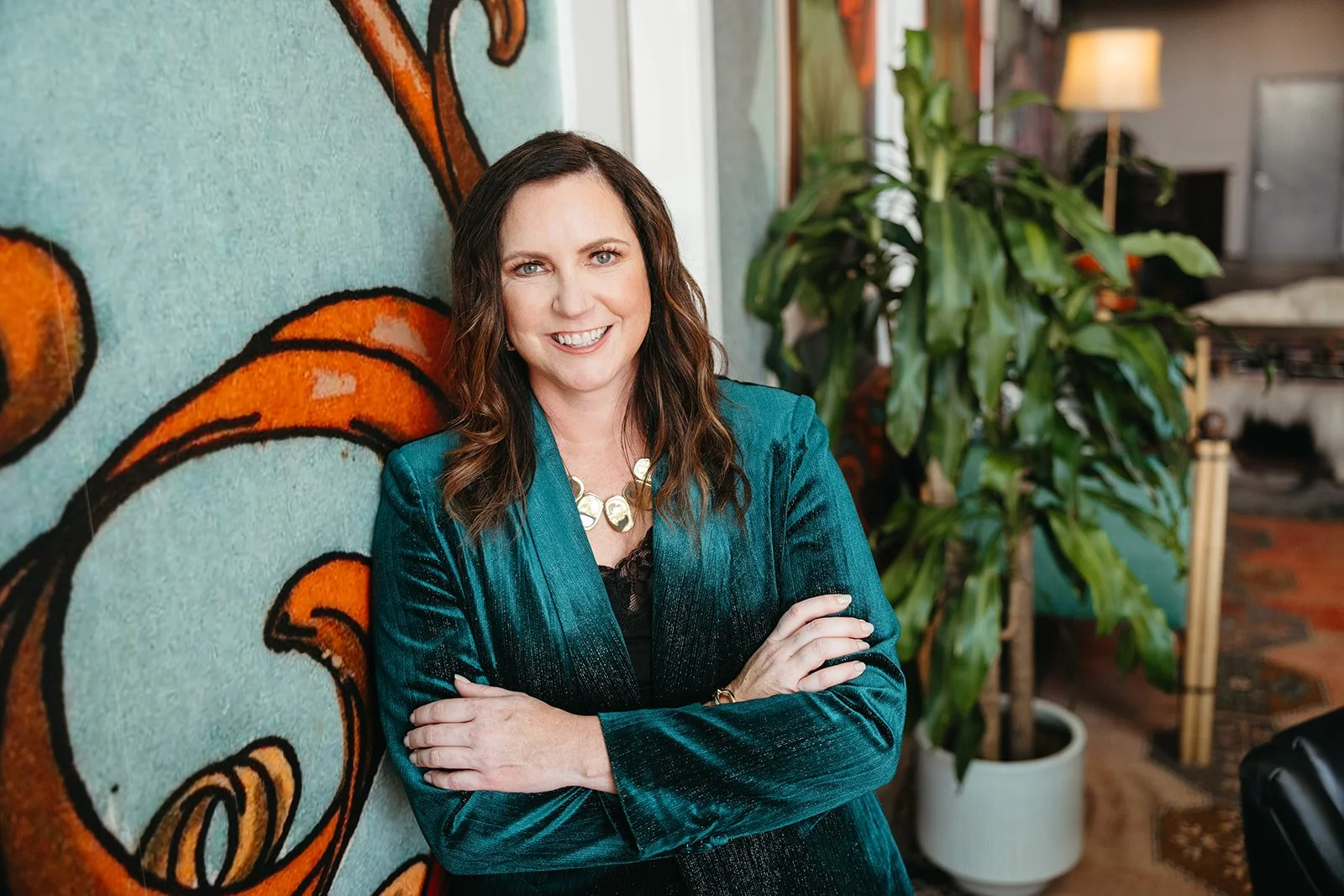 A woman with long brown hair smiling, crossing her arms, standing next to a colorful abstract painting and a large green leafy plant in a warmly lit room.