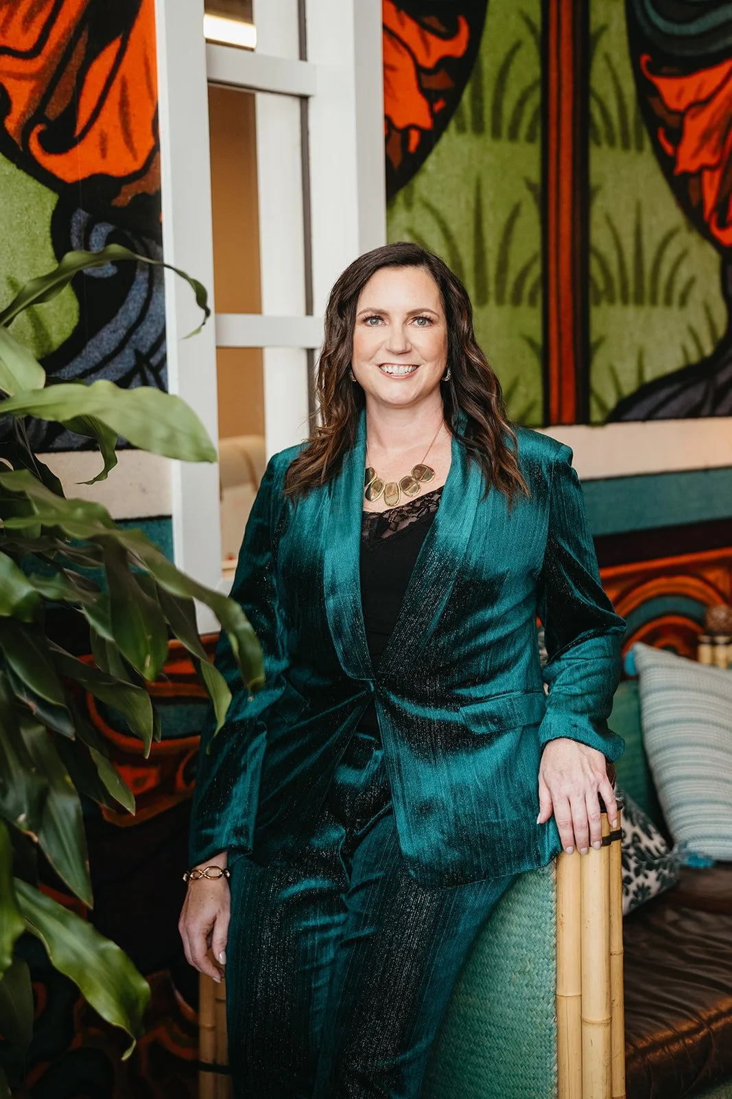 A woman in a teal satin suit standing indoors, smiling, with a colorful abstract mural and green plants in the background.