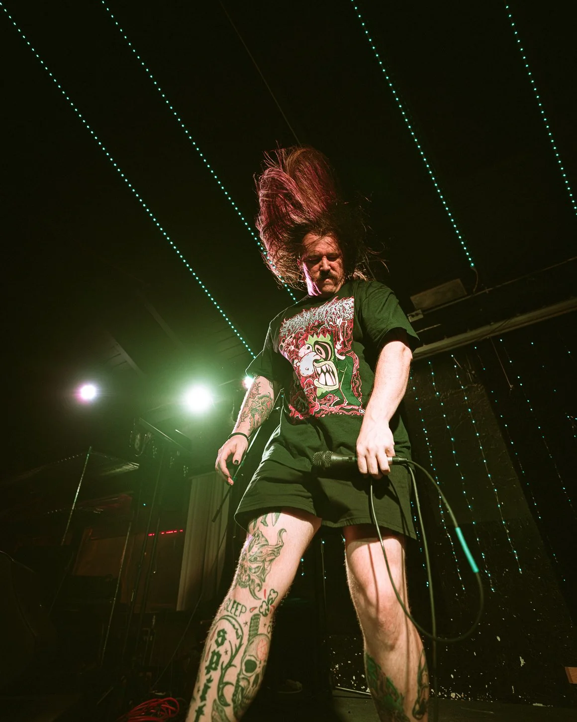 A male musician with long hair, tattoos, wearing a black graphic T-shirt and shorts, performing on stage with a microphone, under colorful stage lighting.