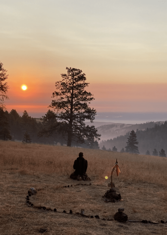 A person sitting on a field at sunset, facing mountains with trees, next to a small memorial with a flag.