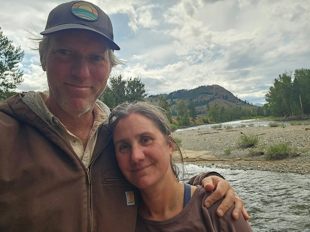 A man and woman outdoors near a river with mountains and trees in the background.