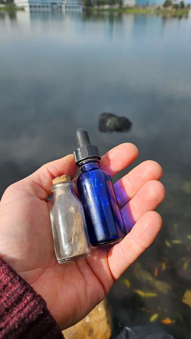 Hand holding two small bottles, one clear with a cork stopper and one dark blue with a dropper, near a body of water with a rock and buildings in the background.