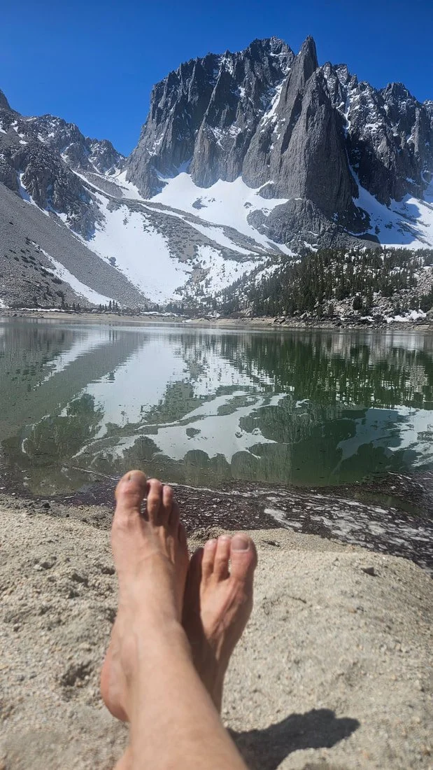 Person relaxing on sandy shore of mountain lake with reflections of snow-capped peaks and forested slopes under a blue sky.