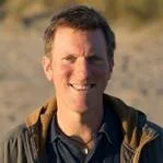 Scott Davidson with short dark hair smiling outdoors, wearing a dark jacket and shirt, with a blurred outdoor background.