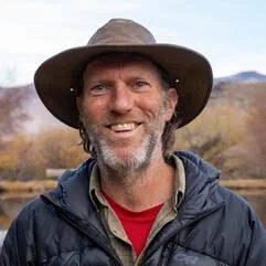Scott Davidson smiling outdoors wearing a wide-brimmed hat, a jacket, and a red shirt, with mountains and trees in the background.