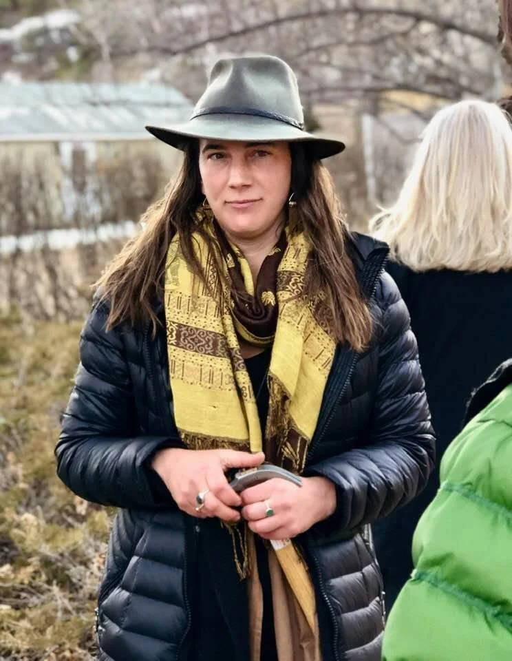 Woman in a gray hat, black puffer jacket, and yellow scarf standing outdoors.