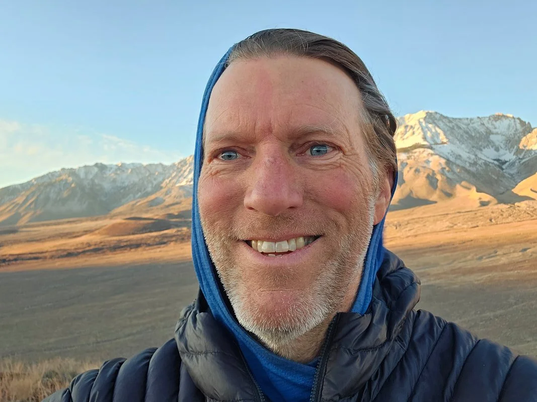 A smiling man outdoors with snow-capped mountains in the background during sunset.