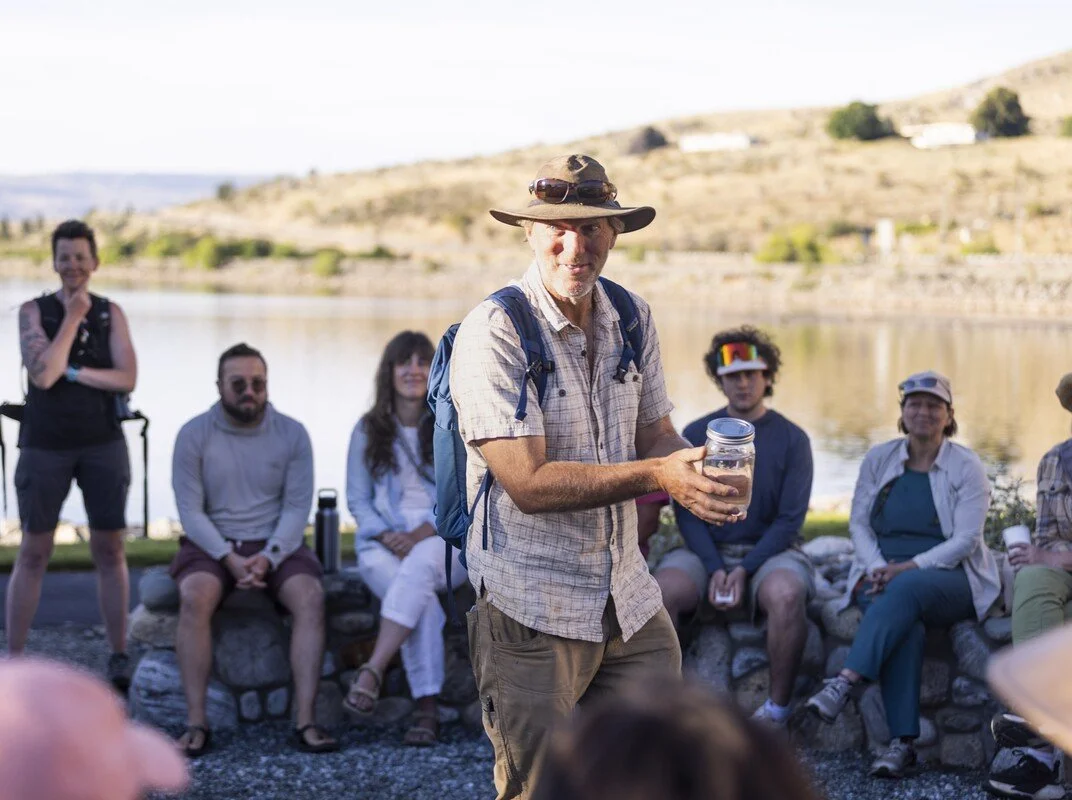 A man with a hat and backpack holding a jar and smiling, standing outdoors near a river with a group of people sitting and standing behind him, in a scenic natural setting with hills in the background.