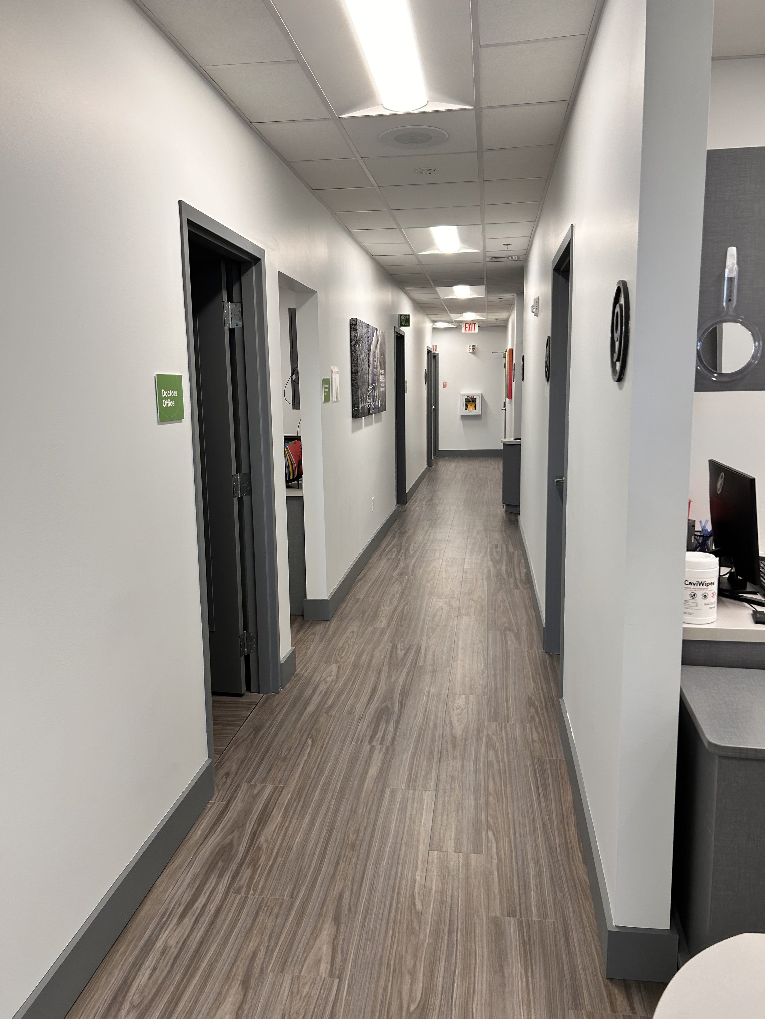 Hospital corridor with multiple doors, signs, and a wood-textured floor, leading to an exit at the end.