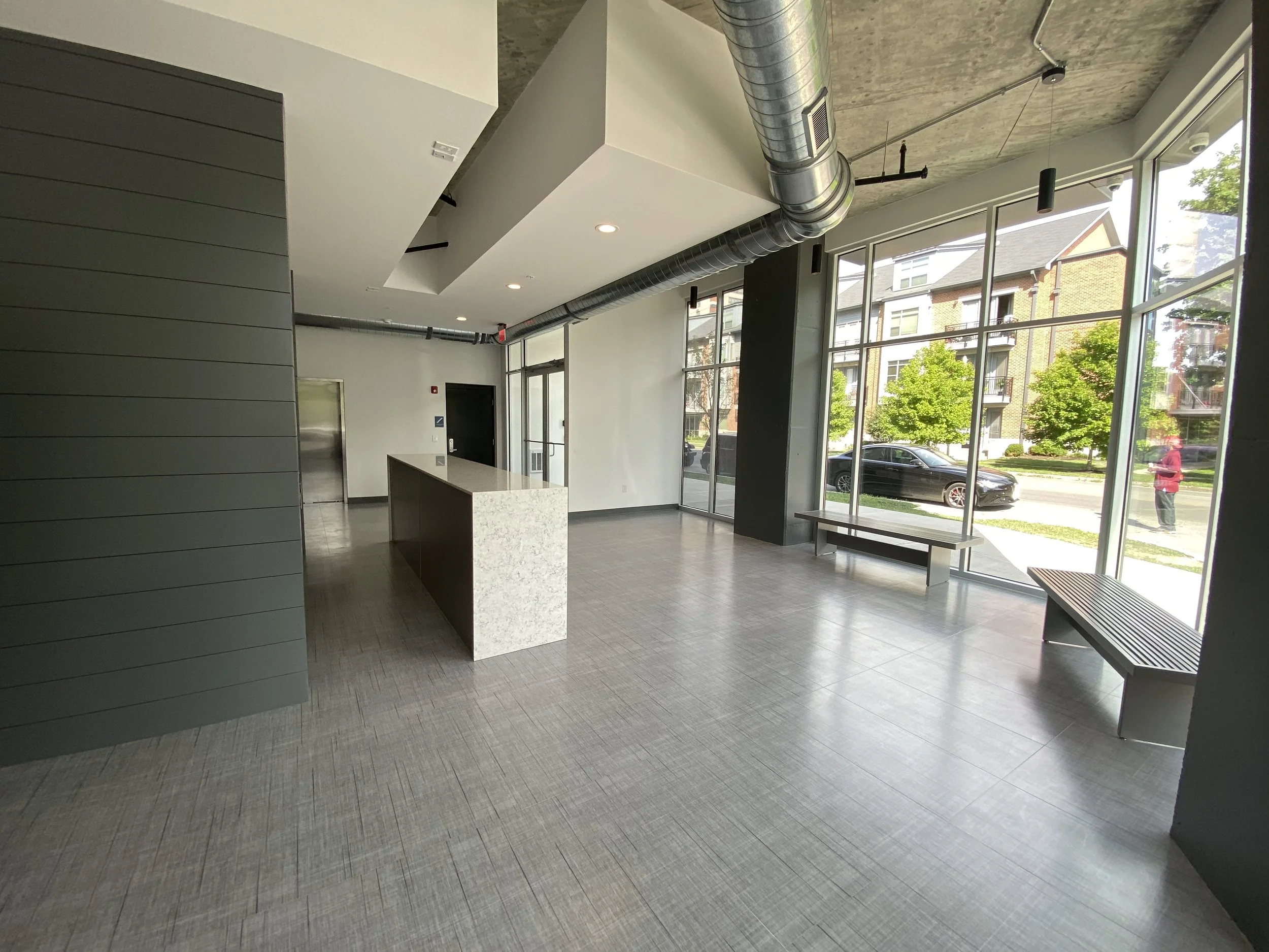 Modern interior space with large windows, gray walls, a white countertop, and benches near the windows. Outside, there are trees and parked cars.