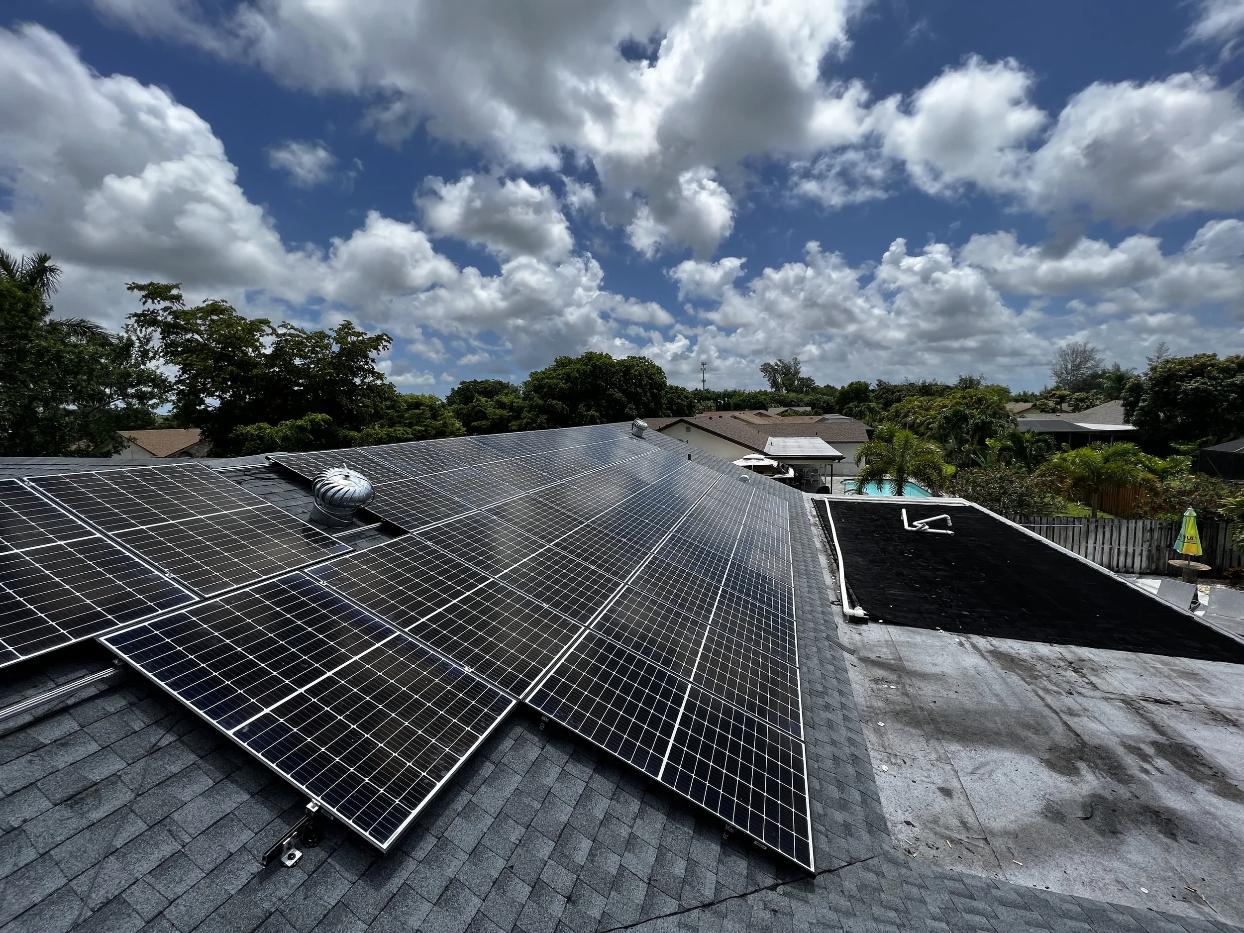 Solar panels installed on a black shingle roof with a cloudy sky and trees in the background.