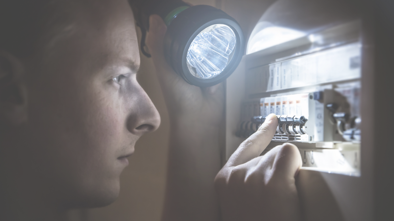A person in a gray hoodie using a flashlight to look at an electrical circuit breaker box.