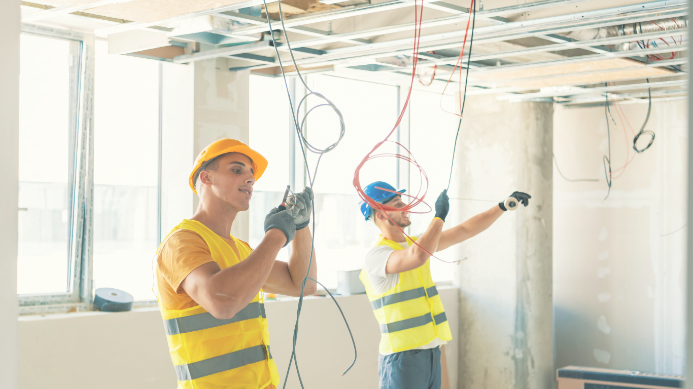 Two construction workers in safety gear working with electrical wires in an unfinished room with large windows.