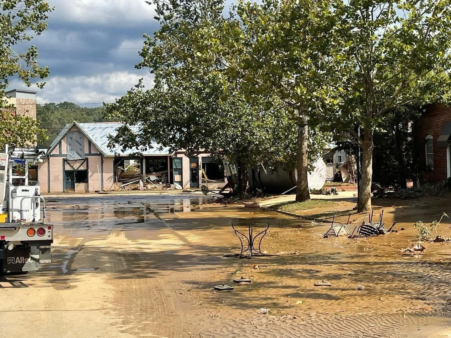 A flooded street with muddy water covering the ground, debris scattered, partially collapsed buildings, and damaged trees during a storm.