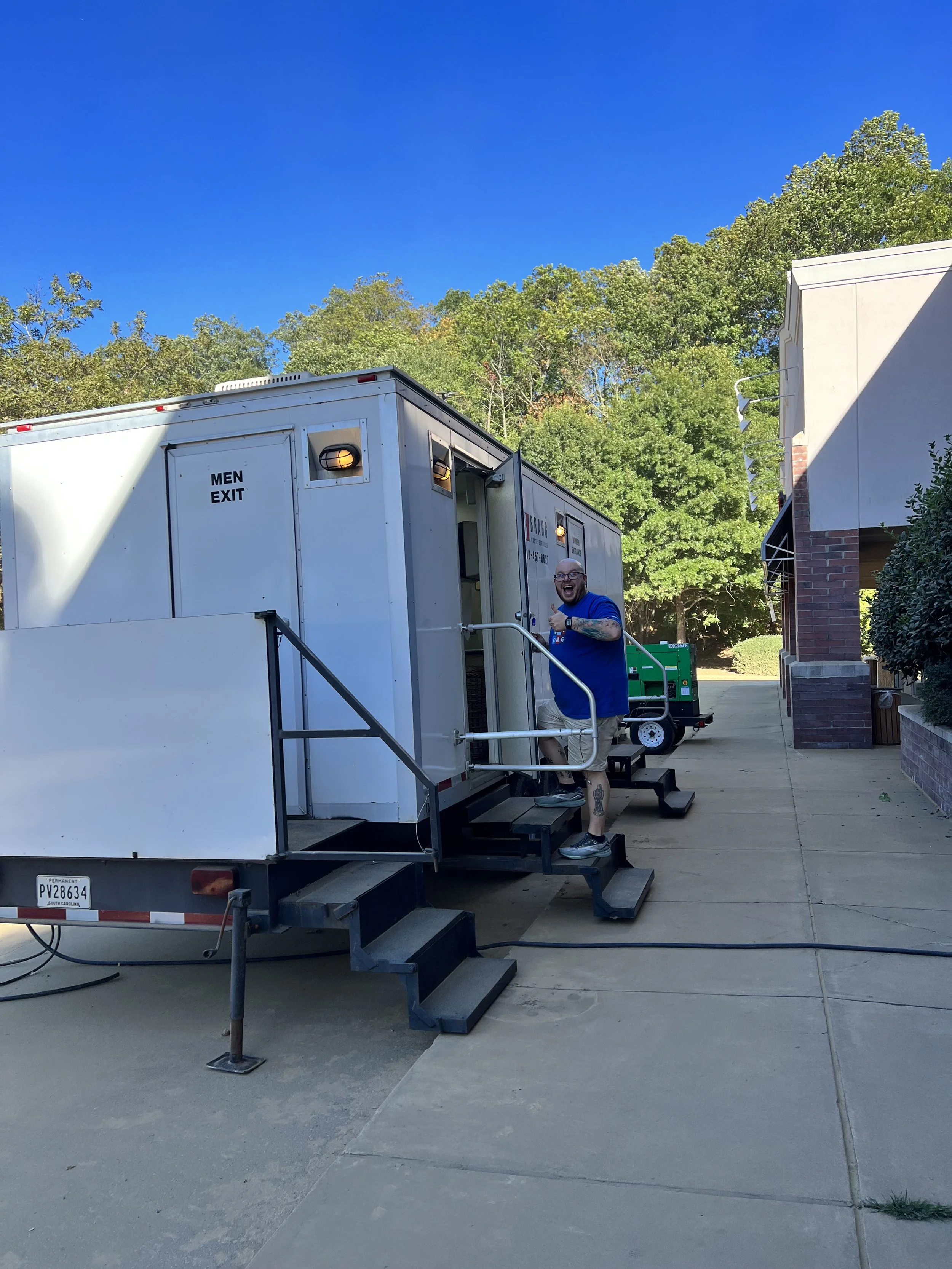 Man smiling and giving a thumbs up while standing on steps outside near a mobile trailer labeled 'MEN EXIT' with a blue sky, green trees, and a building in the background.