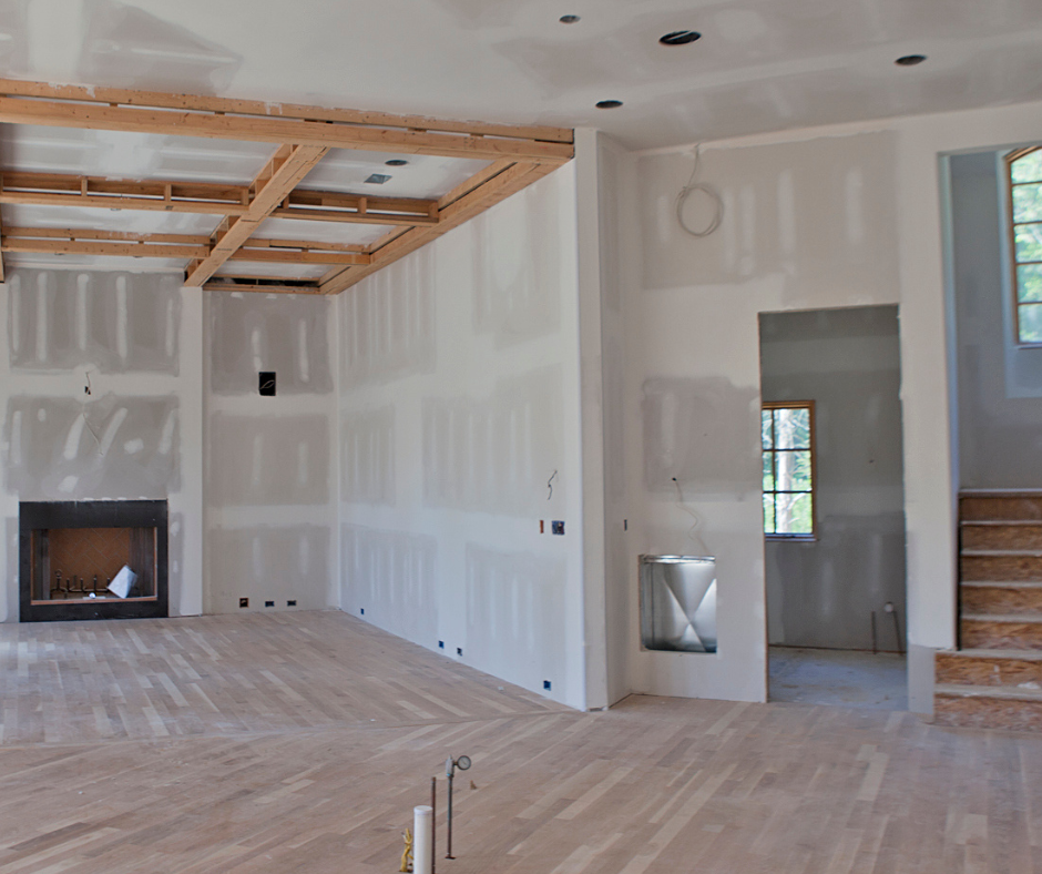 Interior of a house under construction, with drywall partially installed and wooden flooring. Exposed ceiling beams, staircase, and window visible.